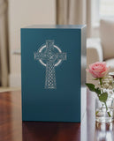 Teal urn with Celtic cross inscribed on a table with a pink rose nearby.