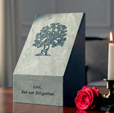 Green slate urn with tree design and 'Lost, but not Forgotten' text, sitting on a table next to a candle and rose.