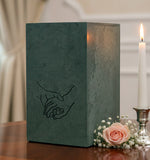 Green Adirondack urn with a line drawing of clasped hands inscribed, placed on a wooden surface with a candle and flowers in the background.
