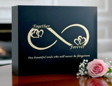 Black Katahdin urn with gold infinity symbol and text, accompanied by a pink rose and baby's breath.