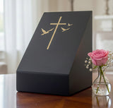 Black slate urn with gold cross design sitting on a table next to a pink rose in a vase.