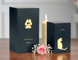 Two black slate pet urns with pawprints and a cat inscribed, a lit candle, and flowers on a table.