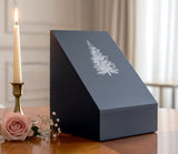 Black slate urn with inscribed tree design set on a table near a candle and flower.