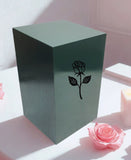 Green painted urn with a rose design on a table next to a pink rose.