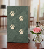 Green slate urn with inscribed paw prints sitting on a table with a candle and flowers in the background