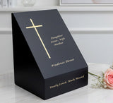 Black slate urn with gold text and cross design sitting on a white surface near a flower.
