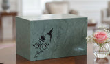 Green Seguin urnwith a hummingbird and flower design inscribed, on a table with a flower near, blurred background