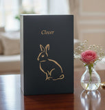 Black slate urn with a gold rabbit inscribed sitting on a table with a pink rose in the background.