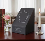 Memorial urn with engraved name and dates on a table with a candle and flowers.